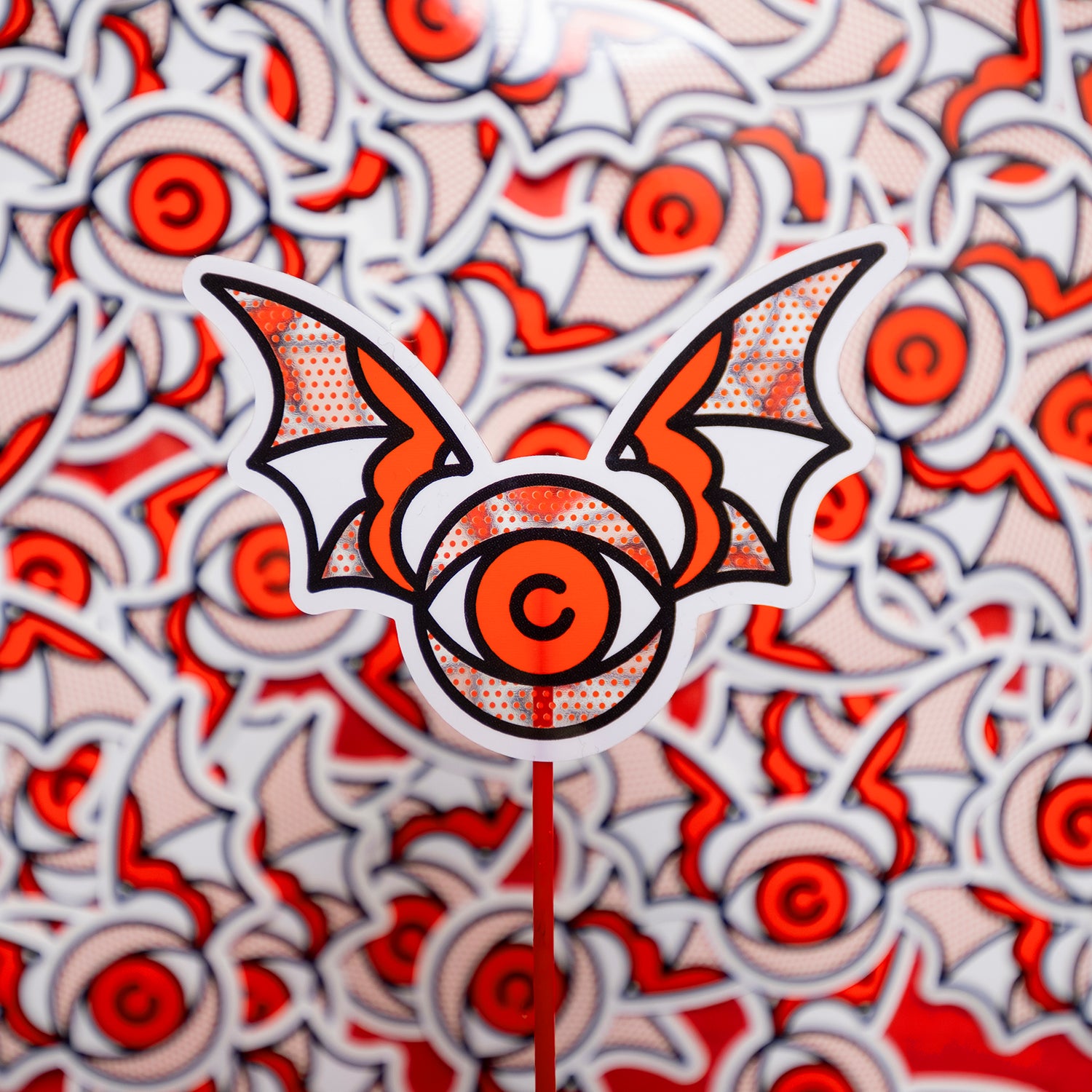 A 3x2.2" clear vinyl art sticker of an eyeball with bat wings drawn in a flash tattoo and pop art fusion style in a red, white and black color palette by the artist, Red Halftone.