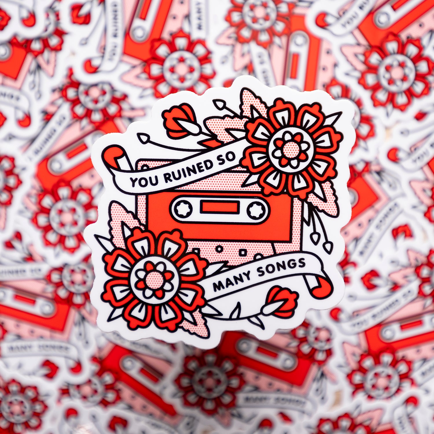 A 3.1x3.1" vinyl art sticker of a cassette tape with two flowers and banners overlaid on adjacent corners with text that reads: You ruined so many songs. Drawn in a flash tattoo and pop art fusion style in a red, white and black color palette by the artist, Red Halftone.